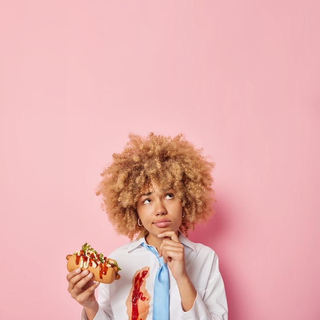 Verticaal schot van peinzende vrouw met krullend haar eet smakelijke hotdog houdt kin geconcentreerd boven het hoofd heeft ongezonde voeding draagt wit overhemd besmeurd met ketchup en blauwe stropdas geïsoleerd over roze muur