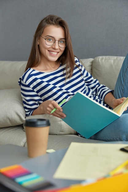 Verticaal schot van vrolijke brunette vrouw controleert cursuswerk, leest tekstinformatie, uit leerboek, houdt pen vast, glimlacht gelukkig, zit op bank drankjes afhaal drank. Hipster-student leert iets