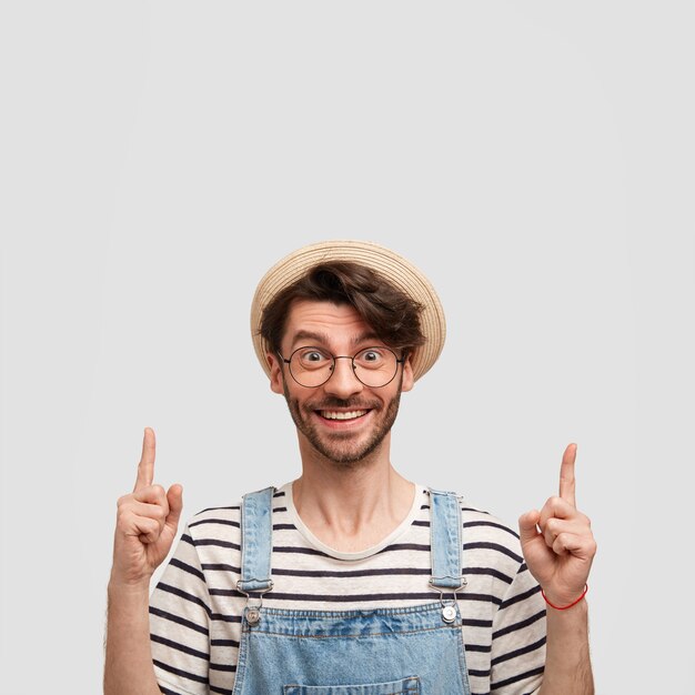 Verticaal schot van vrolijke mannelijke boer met gelukkige uitdrukking, gekleed in casual gestreepte trui en denim tuinbroek, omhoog gericht