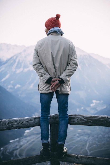 Verticaal shot van een man met een rode hoed en staande op een houten hek met bergen Gratis Foto