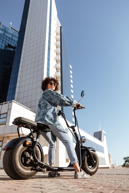 Verticaal zijaanzichtbeeld van onbezorgde krullende vrouw in op moderne motor zitten en zonnebril die in openlucht kijken weg