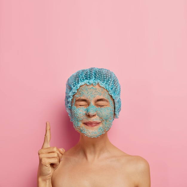 Verticale afbeelding van tevreden schoonheid meisje heeft blauwe zeezout scrub op gezicht, sluit de ogen en wijst wijsvinger naar boven, draagt een badmuts, brengt weekend door in spa salon heeft problematische droge huid, schoonheidsregime