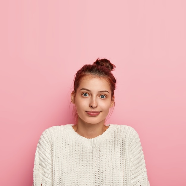 Gratis foto verticale opname van aangenaam ogende tevreden duizendjarige meisje draagt casual gebreide trui, heeft volle lippen en blauwe ogen, donker haar gekamd in knoop, poses op roze achtergrond, lege ruimte erboven