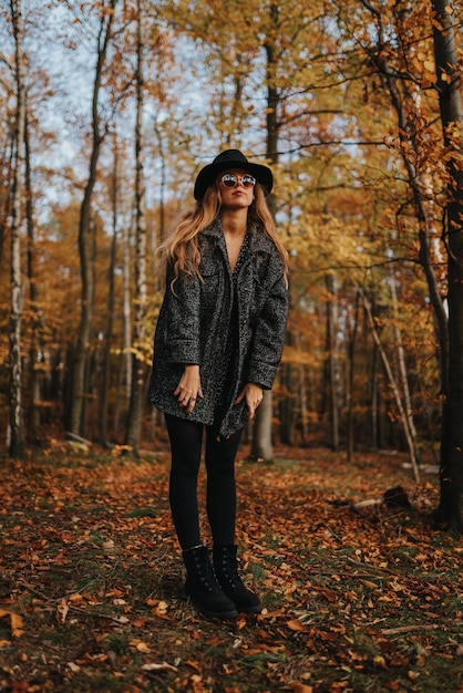 Verticale opname van een blond sexy vrouwelijk model met zwarte jas en hoed poserend in een herfstbos