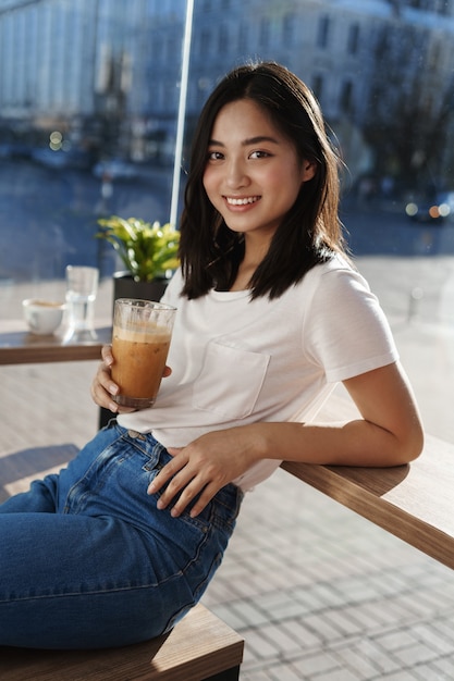 Verticale portret van gelukkig modern meisje, zittend in café in de buurt van raam en leunend op tafel, ijs latte drinken