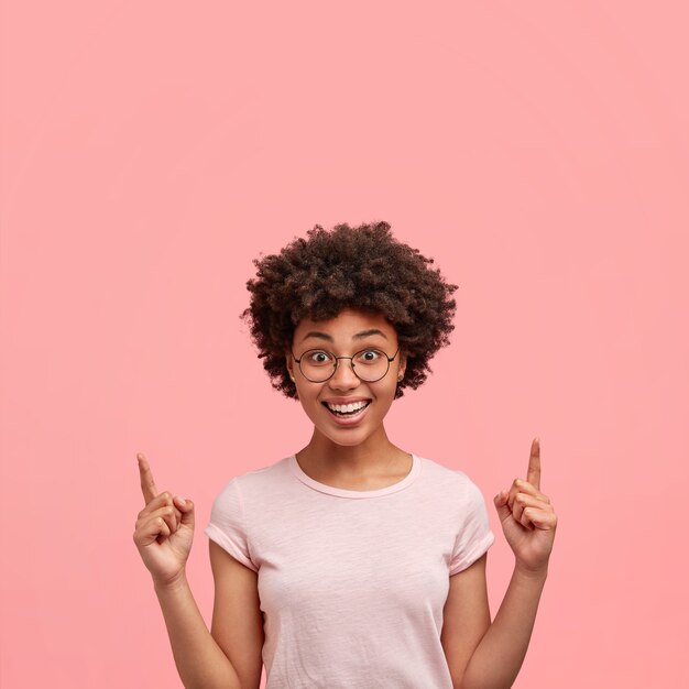 Verticale portret van positieve vrolijke Afro-Amerikaanse vrouw met vriendelijke glimlach, gekleed in vrijetijdskleding, wijst met beide wijsvingers naar boven