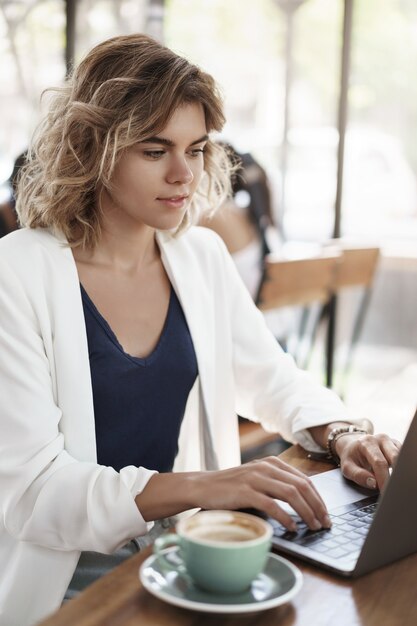 Verticale shot aantrekkelijke succesvolle zakenvrouw stijlvolle witte jas zitten café co-working ruimte koffie drinken cappucino look laptop display typen contact klant, schrijven van rapporten, project voorbereiden.