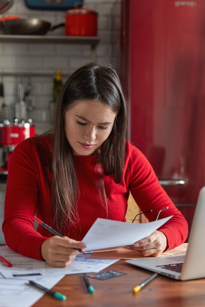 Verticale shot van ernstige sproeten vrouw architect houdt document, krijt, zit aan houten keukentafel, contract bestudeert, toekomstig project voorbereidt, papieren analyseert. Mensen, werk, carrière en baanconcept