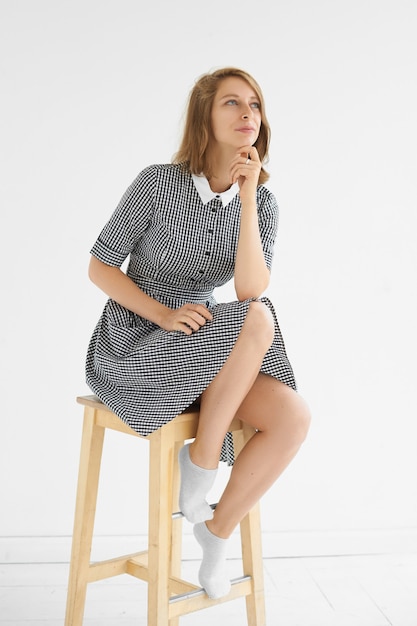 Verticale studio foto van mooie jonge vrouw elegante geruite jurk dragen met witte kraag zittend op hoge houten stoel, met dromerige doordachte blik, haar kin aanraken en glimlachen