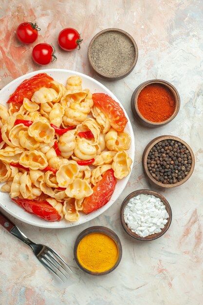 Verticale weergave van smakelijke conchiglie met peper en tomaten op een witte plaat op houten snijplank en mes verschillende kruiden op gemengde kleur achtergrond