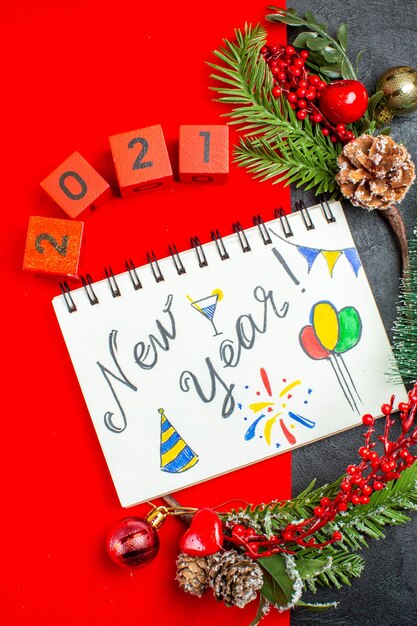 Verticale weergave van spiraal notebook met Nieuwjaar schrijven en tekeningen decoratie accessoires fir takken xsmas sok nummers op een rode servet en kerstboom op donkere achtergrond