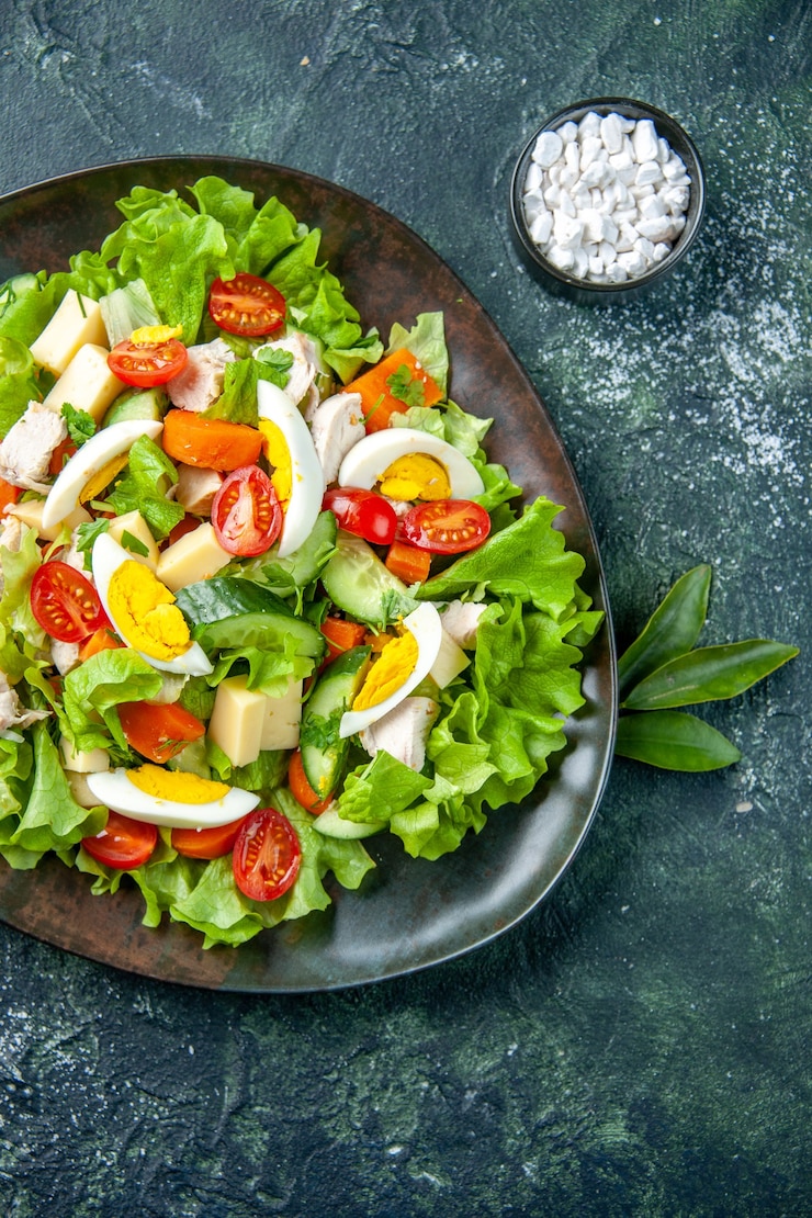 Gratis Foto verticale weergave van zelfgemaakte heerlijke salade met veel ingrediënten in een plaat en zout op zwarte groene mix kleuren achtergrond