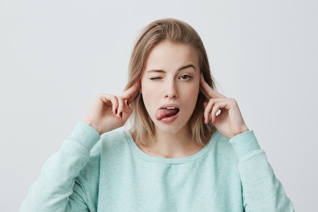 Gratis foto verveeld en geïrriteerd jonge blonde vrouw fronsen en oren dichtknijpen met vingers kan geen geluid verdragen, negeert stressvolle situatie, steekt tong uit. negatieve menselijke emoties