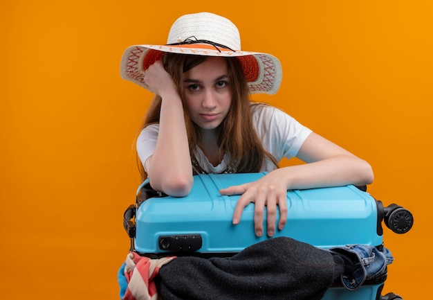 Verveeld jong reizigersmeisje die hoed dragen die koffer vol doeken houden en hand op hoofd zetten op geïsoleerde oranje ruimte met exemplaarruimte