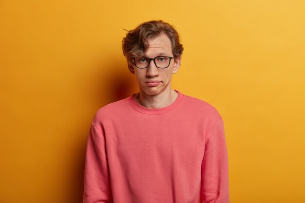 Verveeld, niet onder de indruk mannelijke student kijkt serieus, heeft een uitdrukking van vermoeidheid, draagt een optische bril en een roze trui, zucht van vermoeidheid, geïsoleerd op gele muur. Gezichtsuitdrukkingen