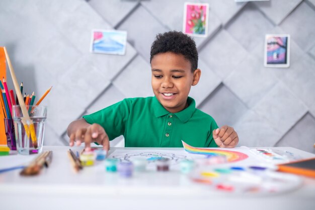 Verven. Lachende donkerhuidige schoolgaande jongen in casual groen t-shirt zittend aan tafel met kunstbenodigdheden die een verftube in lichte kamer aanraken