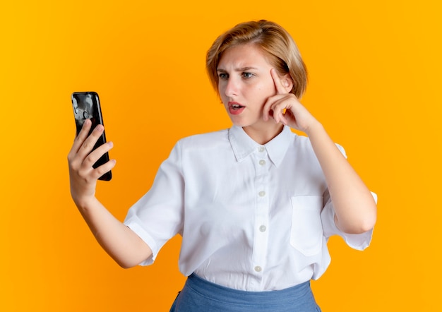verward blond Russisch meisje legt hand op gezicht kijken naar telefoon