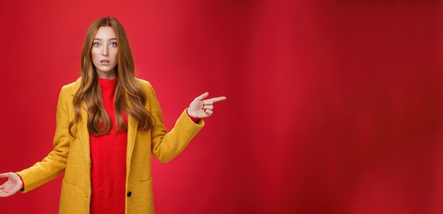 Gratis foto verward en ondervraagd schattig roodharig meisje in gele herfstjas die naar rechts wijst terwijl ze haar schouders ophaalt c