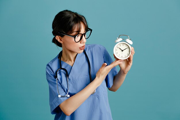 Verward houden wekker jonge vrouwelijke arts dragen uniform fith stethoscoop geïsoleerd op blauwe achtergrond