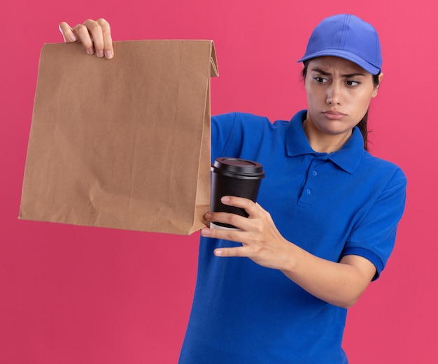 Verward jong bezorgmeisje met uniform met dop die vasthoudt en kijkt naar een papieren voedselpakket met een kopje koffie geïsoleerd op een roze muur