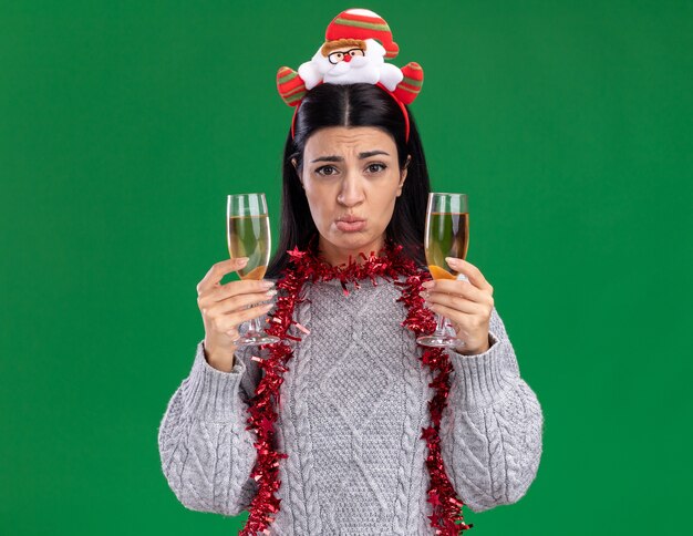 Verward jong Kaukasisch meisje met hoofdband van de kerstman en klatergoud slinger rond nek met twee glazen champagne kijken camera geïsoleerd op groene achtergrond