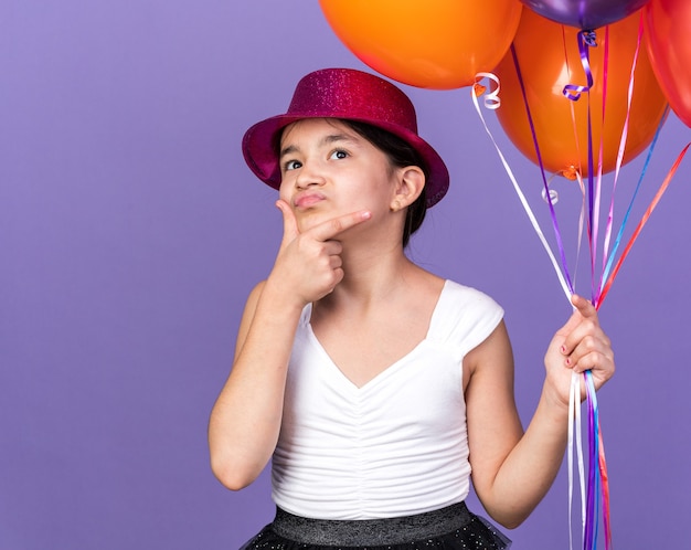 verward jong Kaukasisch meisje met violette feestmuts met helium ballonnen en hand op kin opzoeken geïsoleerd op paarse muur met kopie ruimte