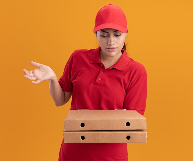 Verward jong leveringsmeisje die eenvormig en GLB dragen die en pizzadozen bekijken die hand verspreiden die op oranje muur wordt geïsoleerd