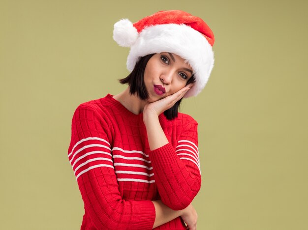 Verward jong meisje met kerstmuts kijken camera hand houden op gezicht met samengeknepen lippen geïsoleerd op olijfgroene achtergrond met kopie ruimte