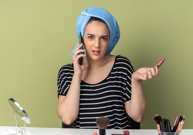 Verward jong mooi meisje zit aan tafel met make-up tools gewikkeld haar in handdoek spreekt op telefoon met lippenstift geïsoleerd op olijfgroene muur