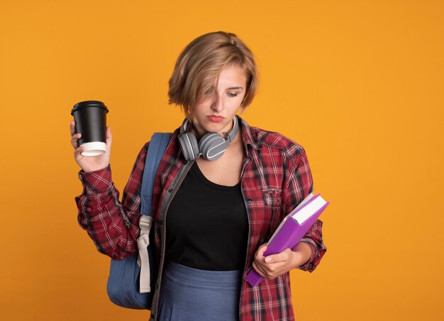 Verward jong slavisch studentenmeisje met koptelefoon met rugzak houdt papieren beker vast en kijkt naar boek en notitieboekje