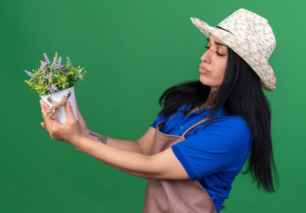 Verward jong tuinmanmeisje met uniform en hoed in profielweergave, vasthoudend en kijkend naar bloempot geïsoleerd op groene muur