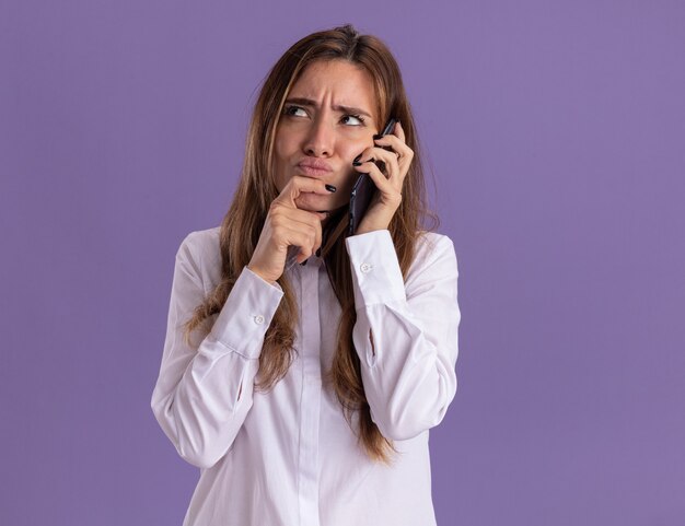 Verward jong, vrij blank meisje praat over de telefoon en legt de hand op de kin en kijkt omhoog geïsoleerd op een paarse muur met kopieerruimte