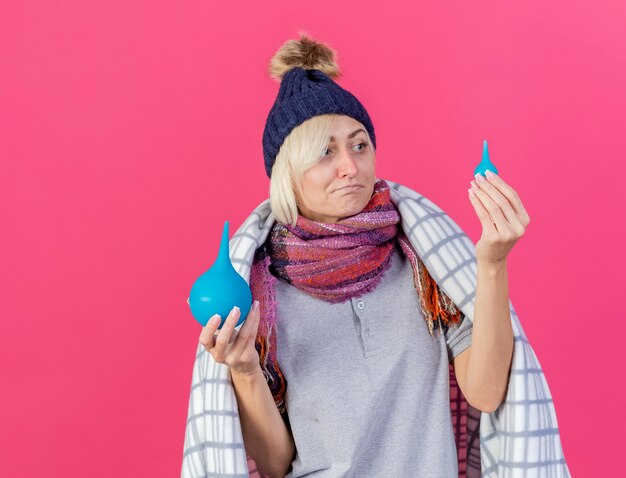 Verward jonge blonde zieke Slavische vrouw met winter muts en sjaal gewikkeld in geruite ruimen en kijkt naar klysma's geïsoleerd op roze muur met kopie ruimte