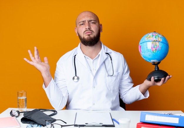 Verward jonge kale mannelijke arts dragen medische gewaad en stethoscoop zit aan bureau met medische hulpmiddelen houden globe en verspreid hand geïsoleerd op een oranje achtergrond