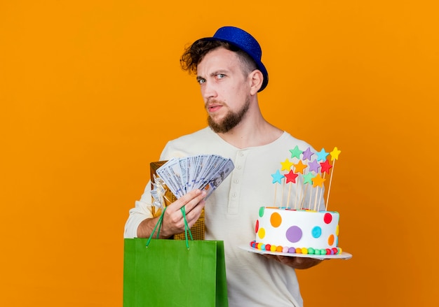 Verward jonge knappe slavische partij kerel met feestmuts bedrijf geschenkdoos geld papieren zak en verjaardagstaart met sterren kijken camera geïsoleerd op een oranje achtergrond met kopie ruimte