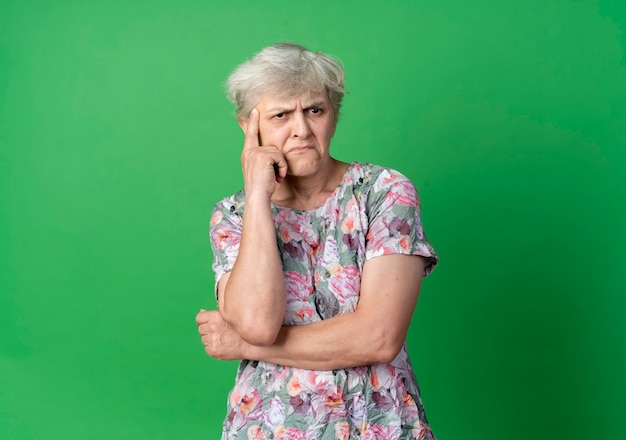 Verward oudere vrouw legt hand op kin kijken kant geïsoleerd op groene muur