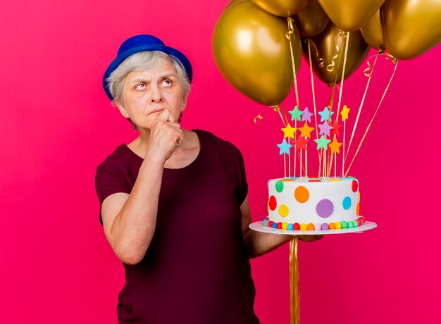 Verward oudere vrouw met feestmuts houdt helium ballonnen en verjaardagstaart zetten hand