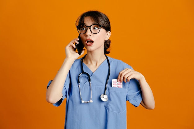 Verward spreekt aan de telefoon jonge vrouwelijke arts met een uniforme stethoscoop geïsoleerd op een oranje achtergrond