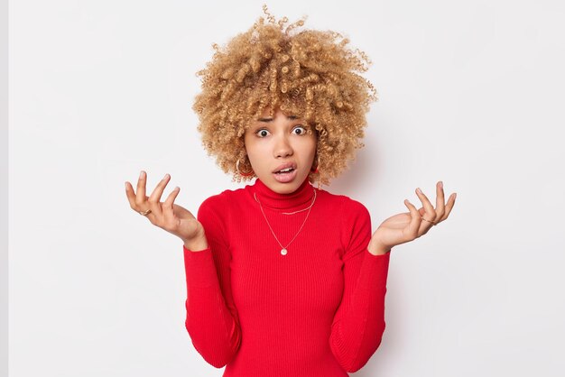 Verward verbaasd bezorgd jonge vrouw met krullend borstelig haar spreidt handpalmen als weet niets haalt schouders op heeft geen idee gezichten moeilijke keuze draagt rode coltrui geïsoleerd op witte achtergrond.