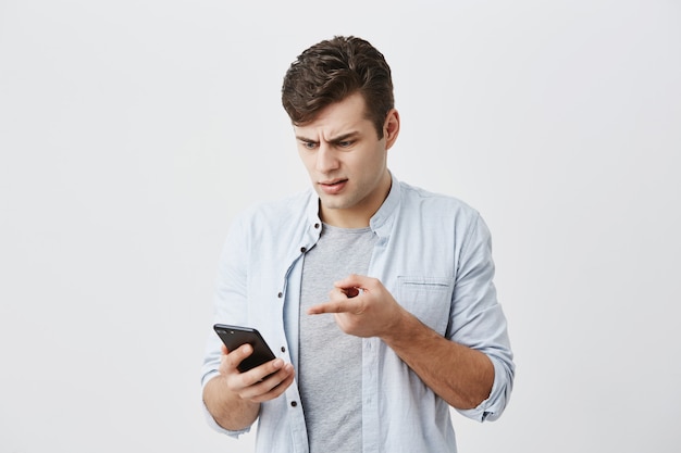 Verward verbaasd mannetje met donker haar, gekleed in een blauw shirt, babbelend en wijzend met de wijsvinger op het scherm van de smartphone, fronsend gezicht in misverstand. Mensen, moderne technologieën