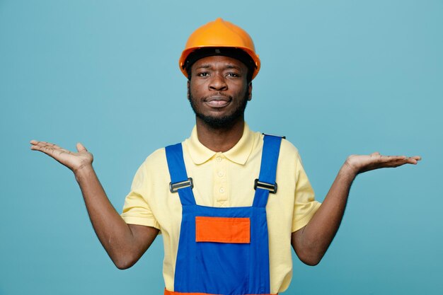 Verward verspreiden handen jonge Afro-Amerikaanse bouwer in uniform geïsoleerd op blauwe achtergrond