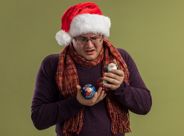 Gratis foto verward volwassen man met bril en kerstmuts met sjaal om nek houden en kijken naar kerstballen geïsoleerd op olijf groene muur