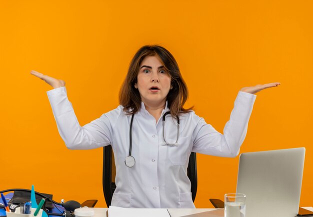 Verward vrouwelijke arts van middelbare leeftijd die medische mantel draagt met een stethoscoop zittend aan een bureau werkt op laptop met medische hulpmiddelen verspreidt handen op geïsoleerde oranje muur met kopie ruimte