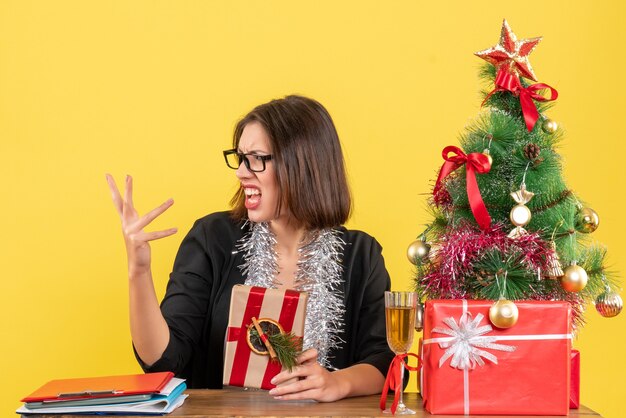 Verward zakelijke dame in pak met bril met haar cadeau en zittend aan een tafel met een kerstboom erop in het kantoor