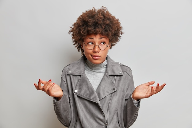 Gratis foto verwarde aarzelende jonge vrouw spreidt handpalmen heeft twijfelachtig gelaatsuitdrukking staat onzeker haalt schouders op