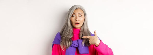 Gratis foto verwarde aziatische senior vrouw met grijs haar hijgend geschrokken wijzend naar zichzelf en kijkend naar camera