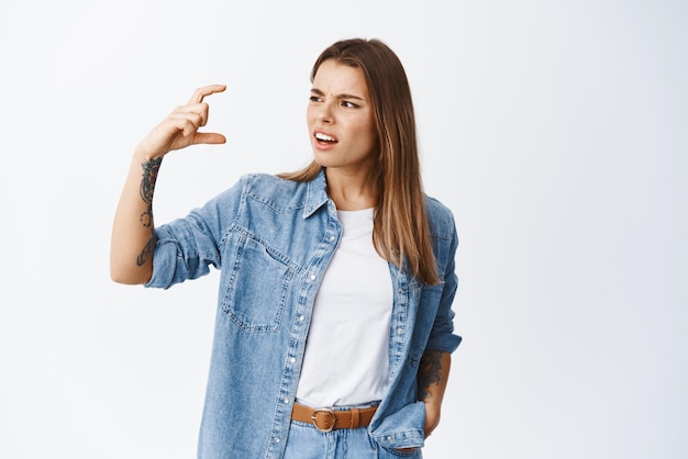 Verwarde en geërgerde jonge vrouw die fronsend naar een klein gebaar staart, teleurgesteld klaagt over een klein ding dat tegen een witte achtergrond staat