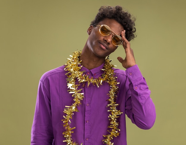 Verwarde jonge Afro-Amerikaanse man met bril met klatergoud slinger rond nek kijken camera aanraken hoofd geïsoleerd op olijfgroene achtergrond