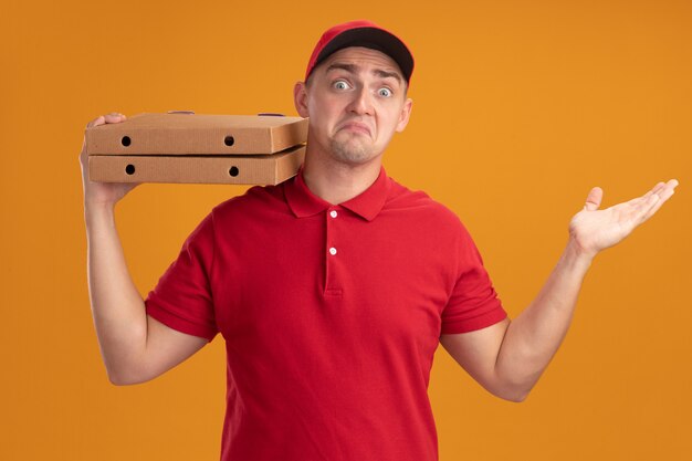 Verwarde jonge bezorger die uniform met pet draagt die pizzadozen op schouder houdt die hand verspreiden die op oranje muur wordt geïsoleerd