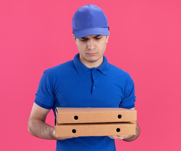 Verwarde jonge blanke bezorger in blauw uniform en pet die pizzapakketten vasthoudt en bekijkt